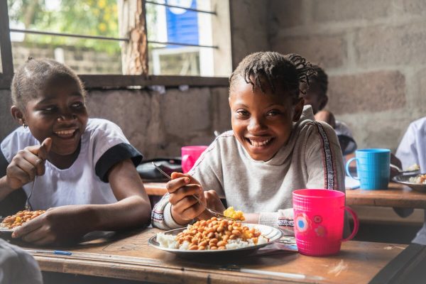 Nsele, province of Kinshasa, Democratic Republic of Congo.
Kamwanya Noëlla (11 years old) eats her meal provided by WFP as part of the school feeding activity. Noëlla is in class of 6eme.

Approximately 13.5 million children in DRC will need humanitarian assistance in 2022, comprising almost 50% of the population in need. School meals help bring and retain children (especially girls) in schools, while contributing to improving their nutritional status, growing the local economy, and reducing poverty when food is locally purchased. In the 2021 – 2022 school year, WFP assisted over 308,000 students in 272 schools.	WFP provides each child with one balanced meal every school day, which is often the only nutritious food they get. WFP is supporting the government in setting up its own school feeding programme in the coming years.  WFP aims to fully support the government’s initiative in school canteens, which targets 3,000 schools in vulnerable areas. For this to succeed, significant resources are to be mobilized while developing more sustainable financing initiatives such as linking schools to revenue-generating farms. School feeding helps to bring and retain children in schools, while contributing to improving their nutritional status, growing the local economy and reducing poverty. WFP plans to reach 500,000 children in the 2023 – 2024 academic year while buying 1200 metric ton of maize and 400 metric ton of beans worth over USD 1 million.